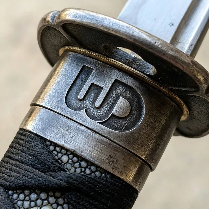 A close-up of a katana hilt featuring a stylized WD logo engraved into the worn, metallic collar (fuchi) just below the handguard.